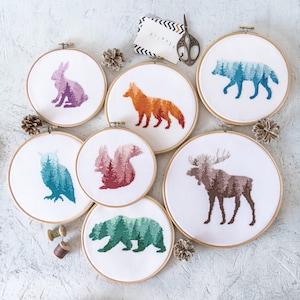 May include: Six embroidery hoops with different animal designs. The designs are a fox, a squirrel, a moose, an owl, a bear, and a wolf. Each animal is made up of a forest scene. The hoops are all white with a wooden frame.