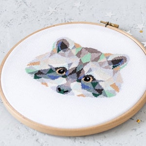 May include: A geometric cross-stitch pattern of a raccoon's face in shades of gray, green, and blue, framed in a wooden embroidery hoop.