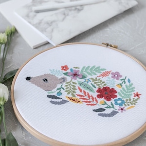 May include: A white embroidery hoop with a cross-stitch design of a hedgehog surrounded by colorful flowers and leaves. The hedgehog is brown and gray with a black nose.