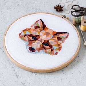May include: A geometric fox cross-stitch pattern in a wooden embroidery hoop. The fox is made up of colorful triangles in shades of orange, brown, pink, blue, and white. The hoop is on a white textured surface.