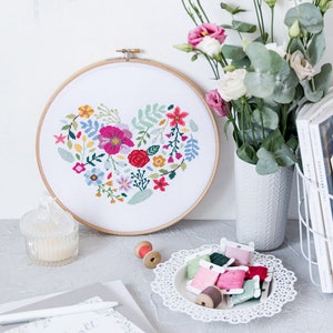K&ouml;nnte beinhalten: Ein wei&szlig;er Stickrahmen mit einem floralen Herzdesign. Der Rahmen ist umgeben von bunten Stickgarn, einer wei&szlig;en Vase mit Blumen und einem wei&szlig;en Drahtkorb mit B&uuml;chern und Kopfh&ouml;rern.