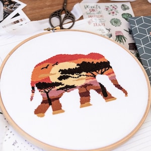 Op de afbeelding: Een borduurring met kruissteek met een kleurrijke zonsondergangsscene in de silhouet van een olifant. De zonsondergang is een gradiënt van oranje, rood en geel met een donkerbruine boom in het midden.