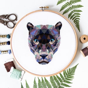 May include: A geometric cross-stitch pattern of a black panther's face with blue eyes. The pattern is stitched on white Aida cloth and framed in a wooden embroidery hoop.