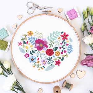 K&ouml;nnte beinhalten: Ein wei&szlig;er Stickrahmen mit einem farbenfrohen Blumenherz-Design. Das Herz besteht aus verschiedenen Blumen, Bl&auml;ttern und Stielen in den Farben Rosa, Rot, Gelb, Blau und Gr&uuml;n. Der Rahmen ist von Holzh Herzen, Stickgarn und Blumen umgeben.