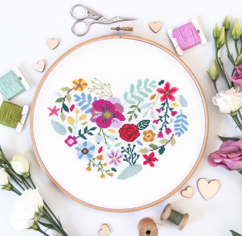 K&ouml;nnte beinhalten: Ein wei&szlig;er Stickrahmen mit einem farbenfrohen Blumenherz-Design. Das Herz besteht aus verschiedenen Blumen, Bl&auml;ttern und Stielen in den Farben Rosa, Rot, Gelb, Blau und Gr&uuml;n. Der Rahmen ist von Holzh Herzen, Stickgarn und Blumen umgeben.