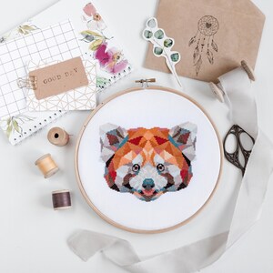 May include: A close-up of a red panda cross-stitch pattern in a wooden embroidery hoop. The red panda is depicted in a geometric style with a colorful palette of red, orange, yellow, blue, and brown.