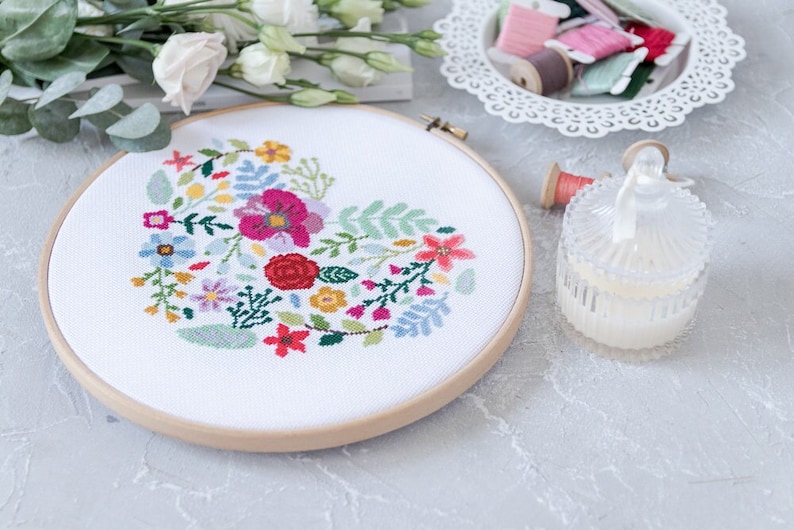 K&ouml;nnte beinhalten: Ein wei&szlig;er Stickrahmen mit einem bunten Blumenmuster in Herzform. Der Rahmen liegt auf einer grauen Oberfl&auml;che mit anderen Bastelutensilien.