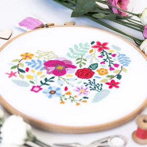 K&ouml;nnte beinhalten: Ein wei&szlig;er Stickrahmen mit einem bunten Blumenmuster in Herzform. Das Design umfasst rosa, rote, gelbe, blaue und gr&uuml;ne Blumen.