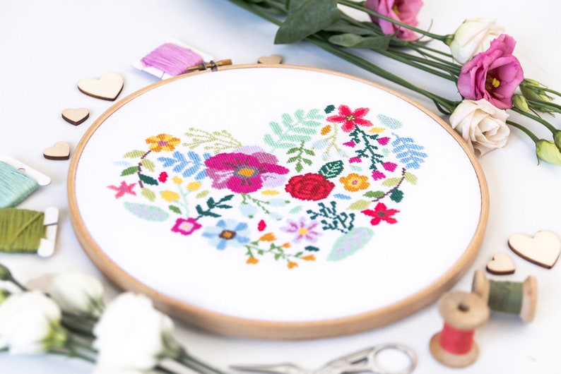 K&ouml;nnte beinhalten: Ein wei&szlig;er Stickrahmen mit einem bunten Blumenmuster in Herzform. Das Design umfasst rosa, rote, gelbe, blaue und gr&uuml;ne Blumen.