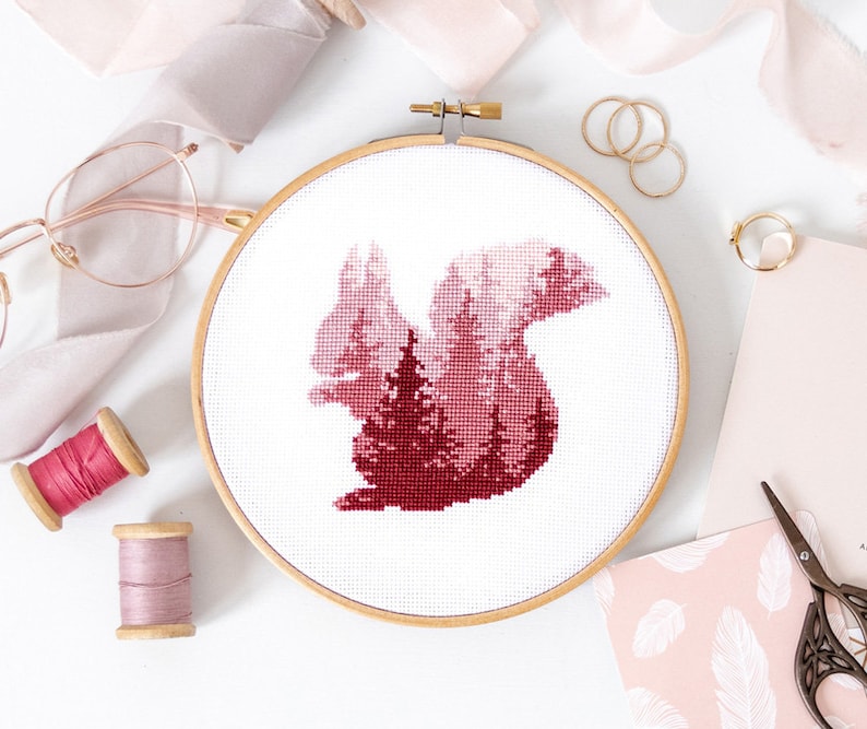 Puede incluir: Un aro de bordado de punto de cruz rojo y blanco con un dise&ntilde;o de silueta de ardilla con un bosque en su interior. El aro est&aacute; sobre una superficie blanca.