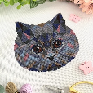 May include: A cross-stitch pattern of a blue cat's face with pink and brown accents. The cat is looking directly at the viewer. The pattern is on white Aida cloth and is surrounded by embroidery floss, scissors, and pink butterfly embellishments.