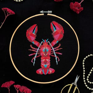 Puede incluir: Un bordado de punto de cruz de una langosta roja con detalles en rosa y azul, sobre un fondo negro dentro de un aro de madera. La imagen incluye tijeras doradas, una guirnalda de cuentas y flores rojas secas.