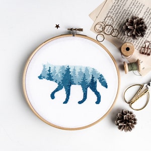 Puede incluir: Una bordado de punto de cruz azul y blanco de una silueta de lobo con un patr&oacute;n de bosque en su interior. El bordado est&aacute; enmarcado en un aro de madera redondo.
