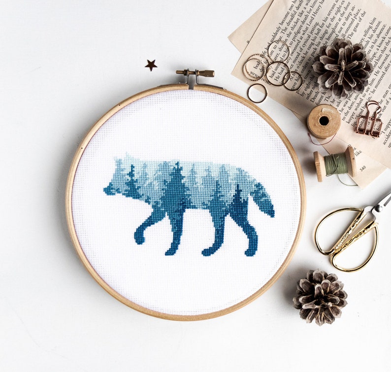 Puede incluir: Una bordado de punto de cruz azul y blanco de una silueta de lobo con un patr&oacute;n de bosque en su interior. El bordado est&aacute; enmarcado en un aro de madera redondo.