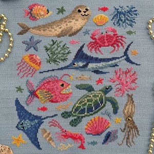 May include: Cross-stitch design featuring various sea creatures, including a seal, fish, crab, and turtle, on a light blue background. The design incorporates coral, starfish, and a stingray. The image is surrounded by seashells, beads, and gold scissors.