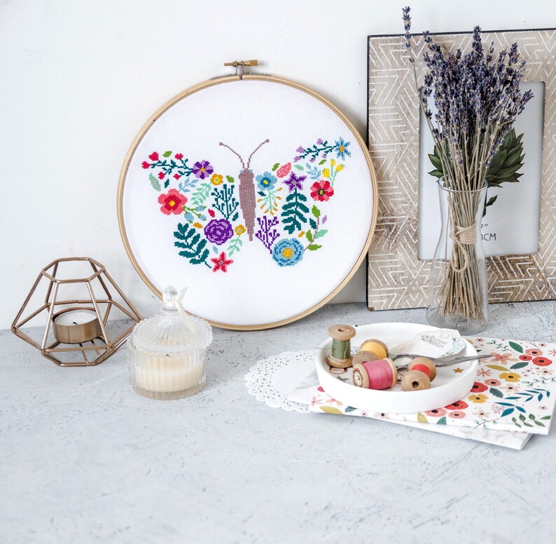 May include: A white embroidery hoop with a colorful butterfly design featuring flowers and leaves. The butterfly is stitched with pink, purple, blue, green, yellow, and red thread. The hoop is on a white surface with a candle, a small plate with spools of thread, and a floral patterned fabric.
