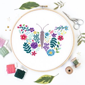 May include: A white embroidery hoop with a colorful floral butterfly design. The butterfly is made up of various flowers and leaves in shades of pink, purple, blue, green, and yellow. The hoop is surrounded by embroidery floss spools and green leaves.