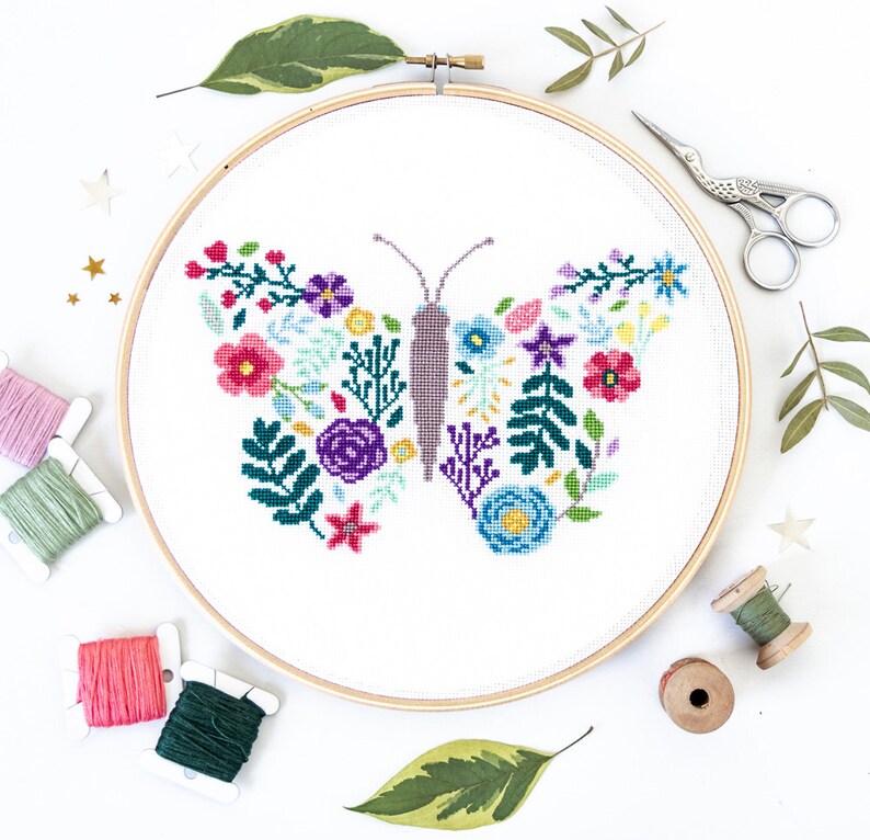 May include: A white embroidery hoop with a colorful floral butterfly design. The butterfly is made up of various flowers and leaves in shades of pink, purple, blue, green, and yellow. The hoop is surrounded by embroidery floss spools and green leaves.