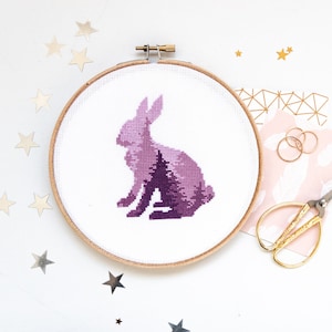 Puede incluir: Un aro de bordado de punto de cruz morado con una silueta de un conejo con un bosque en su interior.