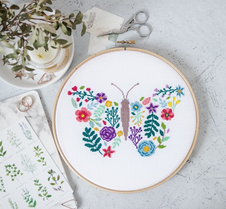 May include: A white embroidery hoop with a colorful floral butterfly design. The butterfly is made up of various flowers and leaves in shades of pink, red, purple, blue, green, and yellow.