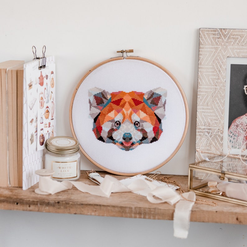 May include: A red panda cross-stitch pattern in a wooden embroidery hoop. The red panda is depicted in a geometric style with colorful, angular shapes.