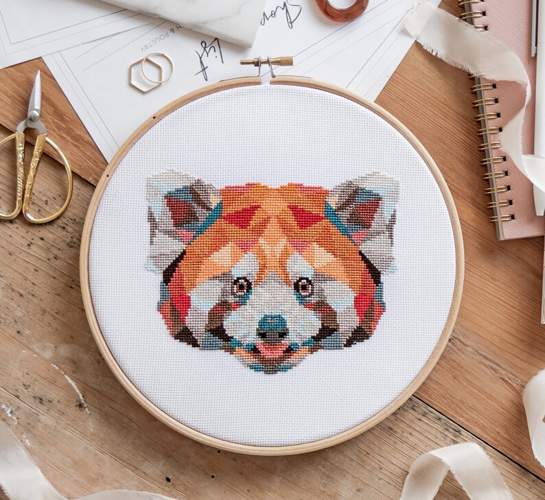 May include: A red panda cross-stitch pattern on white fabric in a wooden embroidery hoop. The red panda is depicted in a colorful, geometric style.