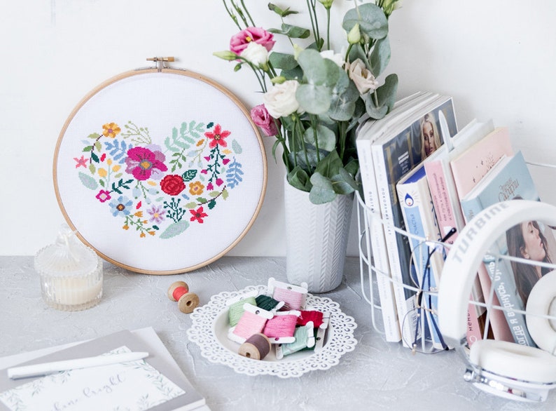 K&ouml;nnte beinhalten: Ein wei&szlig;er Stickrahmen mit einem floralen Herzdesign. Der Rahmen ist umgeben von bunten Stickgarn, einer wei&szlig;en Vase mit Blumen und einem wei&szlig;en Drahtkorb mit B&uuml;chern und Kopfh&ouml;rern.