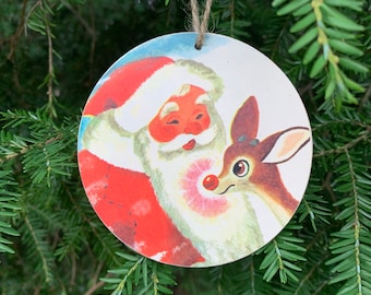 May include: A round, flat, wooden Christmas ornament featuring a vintage-style illustration of Santa Claus with a white beard and red suit, standing next to a brown reindeer with a bright red nose. The ornament hangs from a simple brown twine loop.
