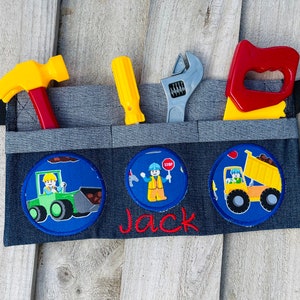 May include: A gray tool belt with a black adjustable strap and pockets for tools. The belt has three embroidered patches with construction workers and vehicles. The name "Jack" is embroidered in red below the patches.