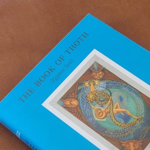 Op de afbeelding: Een blauw boek met de titel "THE BOOK OF THOTH" met "(Egyptian Tarot)" onder de titel. De omslag toont een illustratie binnen een witte rand, die een figuur met een slang in een cirkelvormig ontwerp afbeeldt. Het boek ligt op een bruin oppervlak.