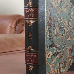 May include: A green leather-bound book with gold accents and marbled paper cover. The title "JOHN DEE" is printed in gold on a burgundy background. The author "CHARLOTTE FELL SMITH" is printed in gold on a burgundy background.