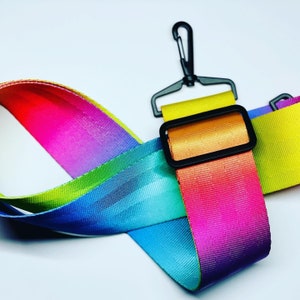 May include: A rainbow-colored adjustable camera strap with a black metal clip and a black adjustable buckle. The strap is made of a woven nylon material.