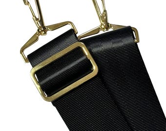 May include: A black nylon bag strap featuring gold-toned metal hardware, including two swivel lobster claw clasps and a rectangular sliding buckle for length adjustment. The strap is made of a durable, woven fabric with a smooth texture.