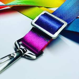 Puede incluir: Un cordón de rayas arcoíris con un clip plateado y una hebilla plateada. La hebilla es de forma rectangular con esquinas redondeadas y tiene una correa morada.