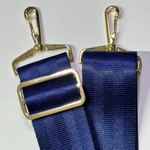 May include: Close-up of two navy blue adjustable straps with gold-colored metal hardware. Each strap has a gold-colored metal clasp and a gold-colored metal rectangular buckle. The straps are made of a woven material.