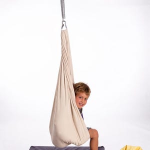 Boy sitting in cream color restNest sensory swing chair with spring suspension, indoor hammock swing, Montessori-inspired kids room decor made from soft cotton fabric for sensory regulation and cosy relaxation.