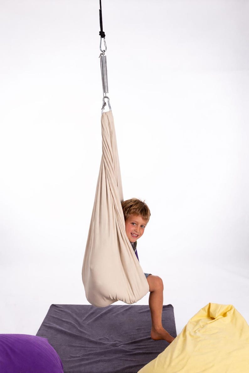 Boy sitting in cream color restNest sensory swing chair with spring suspension, indoor hammock swing, Montessori-inspired kids room decor made from soft cotton fabric for sensory regulation and cosy relaxation.