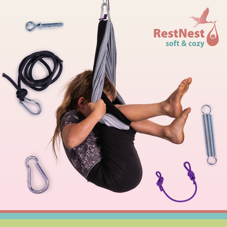K&ouml;nnte beinhalten: Ein Kind h&auml;ngt in einer schwarzen und grauen Stoffschaukel, mit dem Text "RestNest soft & cozy". Das Bild enth&auml;lt eine Feder, Karabinerhaken, ein Seil und eine lila Schleife. Der Hintergrund ist hellrosa mit horizontalen Streifen unten.