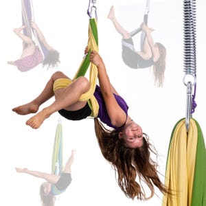 K&ouml;nnte beinhalten: Eine junge Person h&auml;ngt kopf&uuml;ber in einer gelb-gr&uuml;nen Aerial-Yoga-Schaukel. Die Schaukel ist an einer Metallfeder und einem silbernen Karabiner befestigt. Weitere Aerial-Yoga-Schaukeln sind im Hintergrund zu sehen.