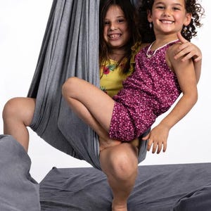 Girls sitting in a grey restNest sensory swing chair with spring suspension, indoor hammock swing, Montessori-inspired kids room decor made from soft cotton fabric for sensory regulation and cosy relaxation.