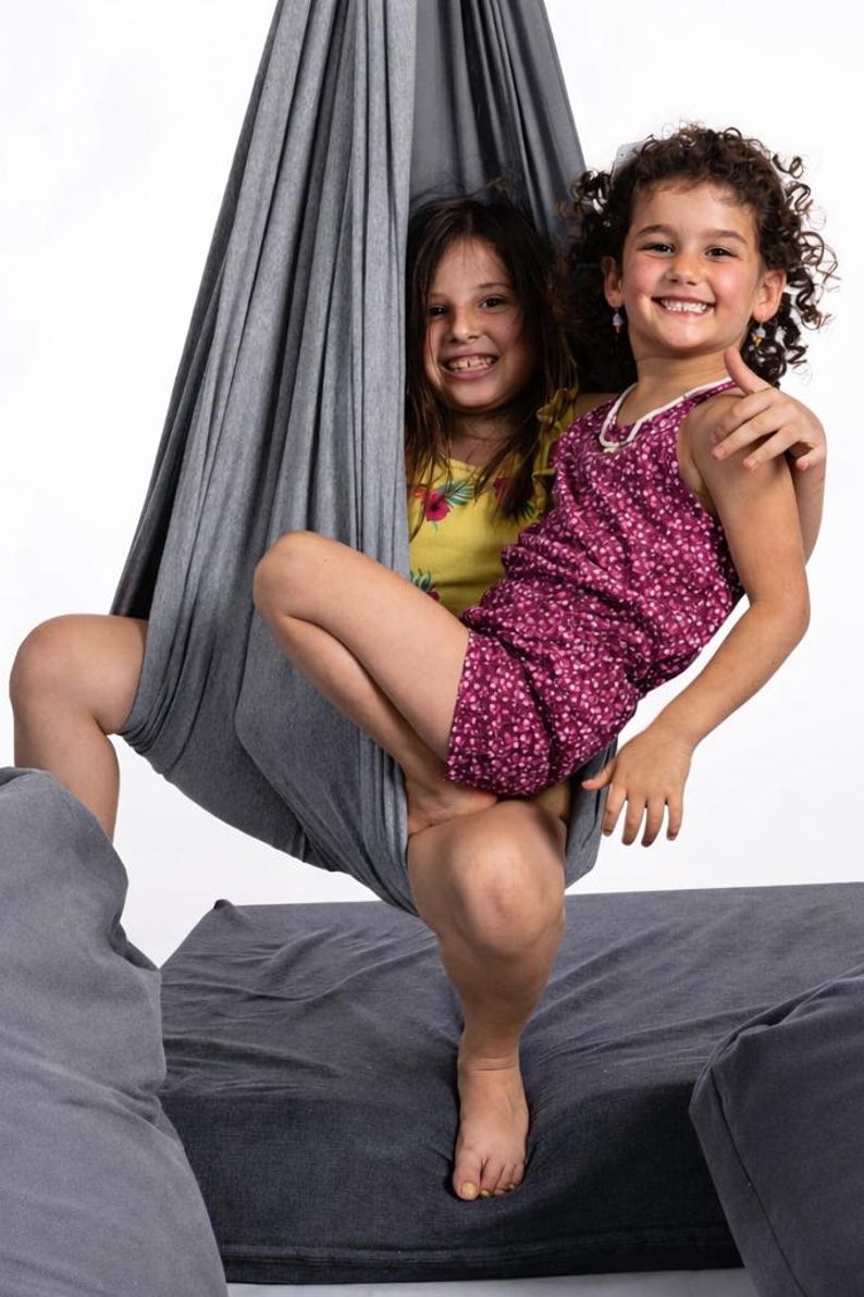 Girls sitting in a grey restNest sensory swing chair with spring suspension, indoor hammock swing, Montessori-inspired kids room decor made from soft cotton fabric for sensory regulation and cosy relaxation.
