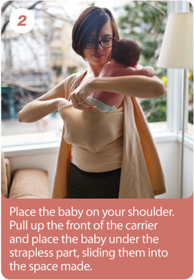 May include: A woman wearing a beige baby carrier, holding a baby. The carrier is a soft, wrap-style design. The image includes text that reads: "Place the baby on your shoulder. Pull up the front of the carrier and place the baby under the strapless part, sliding them into the space made."