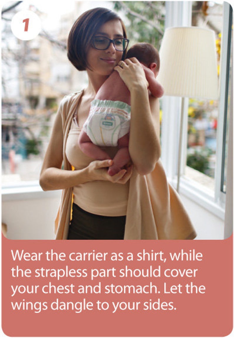 May include: A woman holding a baby wearing a diaper. The woman is wearing glasses and a beige top. The baby is being held in front of a window. The image includes text that reads, "Wear the carrier as a shirt, while the strapless part should cover your chest and stomach. Let the wings dangle to your sides."
