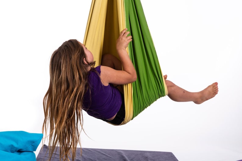 Puede incluir: Un ni&ntilde;o con una camiseta morada y pantalones cortos negros est&aacute; suspendido en un columpio de tela verde y amarilla. El columpio cuelga de arriba y el ni&ntilde;o mira hacia arriba. El fondo es blanco.