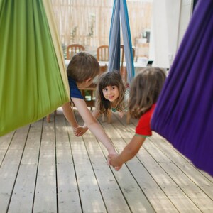 K&ouml;nnte beinhalten: Drei Kinder spielen in bunten H&auml;nge-H&auml;ngematten. Die H&auml;ngematten sind in Gr&uuml;n-, Blau- und Lilat&ouml;nen gehalten. Die Kinder halten sich an den H&auml;nden und l&auml;cheln, was eine spielerische Szene auf einer Holzterrasse ergibt.
