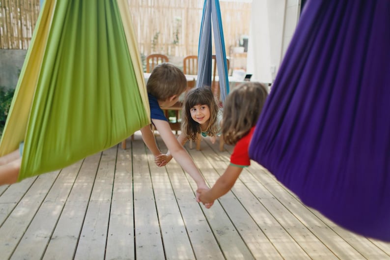 K&ouml;nnte beinhalten: Drei Kinder spielen in bunten H&auml;nge-H&auml;ngematten. Die H&auml;ngematten sind in Gr&uuml;n-, Blau- und Lilat&ouml;nen gehalten. Die Kinder halten sich an den H&auml;nden und l&auml;cheln, was eine spielerische Szene auf einer Holzterrasse ergibt.