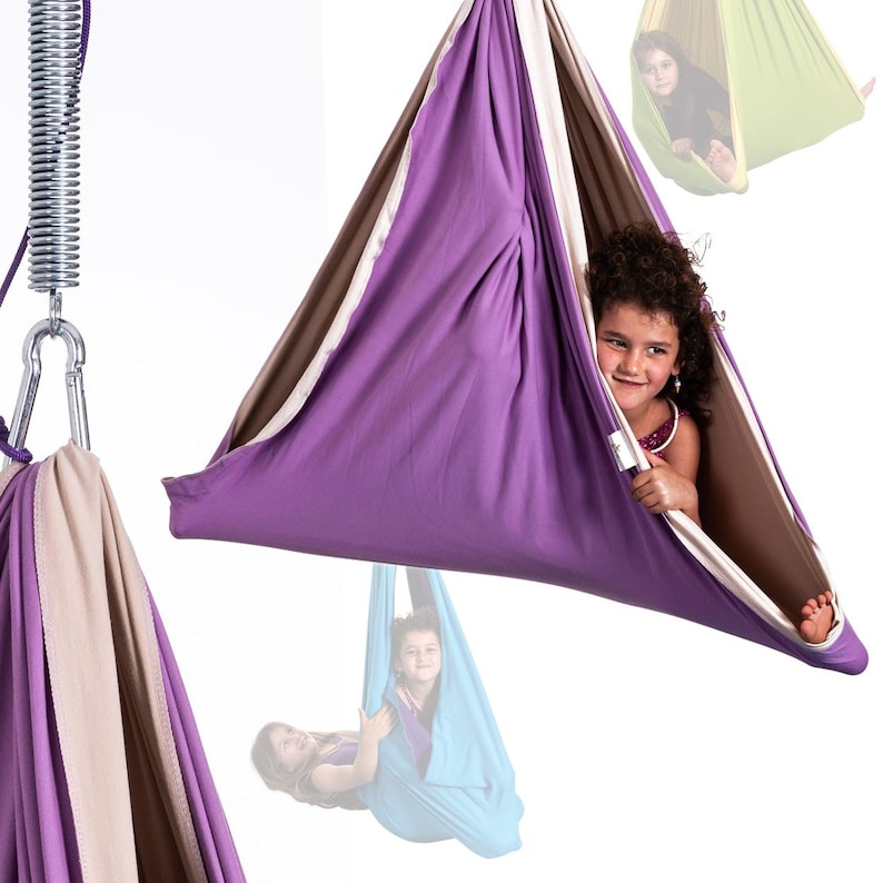 K&ouml;nnte beinhalten: Eine violette, braune und beige sensorische Schaukel mit einem Kind darin. Die Schaukel ist an einer Metallfeder und einem Karabiner befestigt. Weitere Schaukeln in Gr&uuml;n und Blau sind im Hintergrund zu sehen.