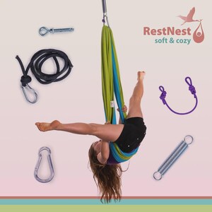 Puede incluir: Una ni&ntilde;a est&aacute; suspendida boca abajo en una hamaca de yoga a&eacute;rea verde y azul. La imagen incluye varios accesorios: un mosquet&oacute;n, un resorte, una cuerda con un mosquet&oacute;n y un lazo de cuerda morada. El texto "RestNest soft & cozy" es visible.