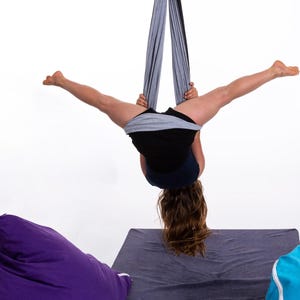 Puede incluir: Una persona practicando yoga a&eacute;reo, suspendida boca abajo de una tela gris. La persona lleva pantalones cortos negros y una camiseta negra. El fondo es blanco, con pufs morados y azules visibles.