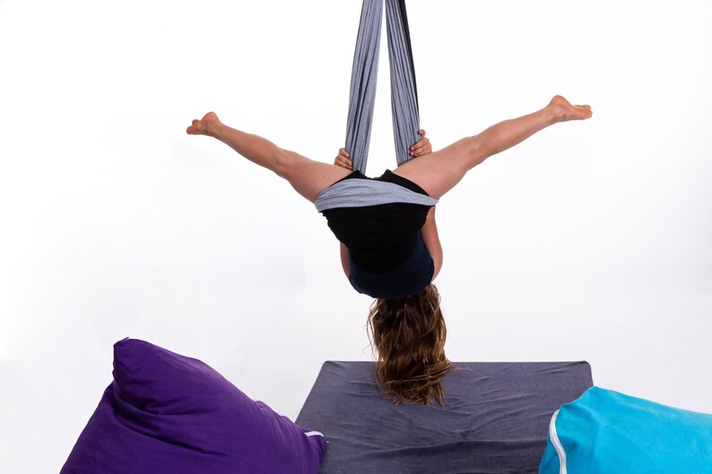 Puede incluir: Una persona practicando yoga a&eacute;reo, suspendida boca abajo de una tela gris. La persona lleva pantalones cortos negros y una camiseta negra. El fondo es blanco, con pufs morados y azules visibles.